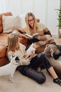 A cozy family scene with mother and daughter reading books together on the sofa with their dogs.