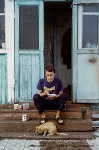 A man sits on steps eating, with a cat beside him, outside a rustic house with teal doors.
