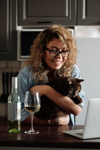 Smiling woman holding a cat while drinking wine and using a laptop for a video call.