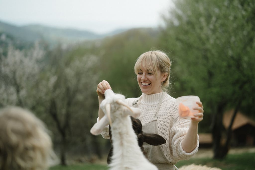 A smiling woman feeds a goat outdoors, capturing a peaceful countryside moment.